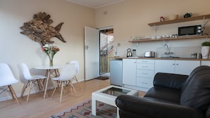 A modern living room with a black leather sofa, a white coffee table, and a dining area with white chairs and a small table. A kitchen with white cabinets and a microwave is visible in the background.