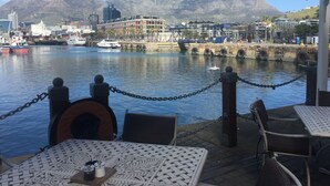 A waterfront dining area with tables and chairs, a view of the harbor, boats, and buildings in the background.