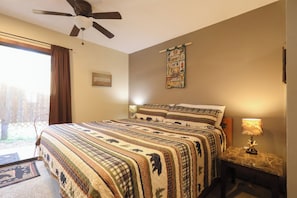 A bedroom with a bed, a ceiling fan, a wall tapestry, a nightstand with a lamp, and a window with curtains.