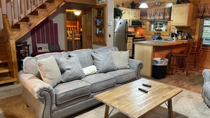 A cozy cabin interior with a wooden staircase, a grey sofa, a wooden coffee table, and a kitchen area with wooden cabinets and a refrigerator. - Cutest, comfiest log cabin with great screened in porch! (Sevierville)