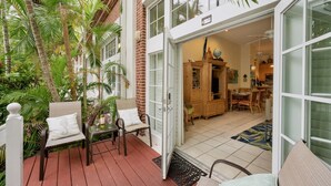 A covered patio with a wooden table and chairs, surrounded by lush greenery and a brick wall.