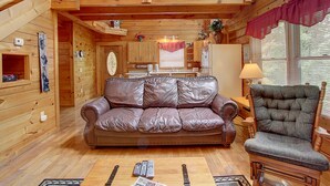 A wooden cabin interior with a leather sofa, a wooden rocking chair, a kitchen area, and a balcony with wooden railings.