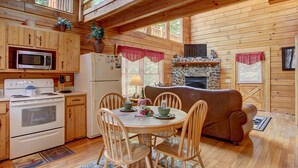 A wooden cabin with a kitchen featuring a stove, microwave, refrigerator, and a stone fireplace. There is a dining table with chairs and a couch in the living area.