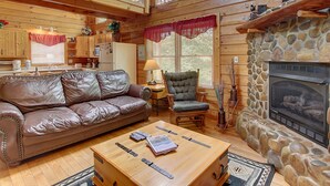 A cabin interior with a wooden floor, leather sofa, a coffee table, a stone fireplace, and a kitchen area with wooden cabinets and a refrigerator.
