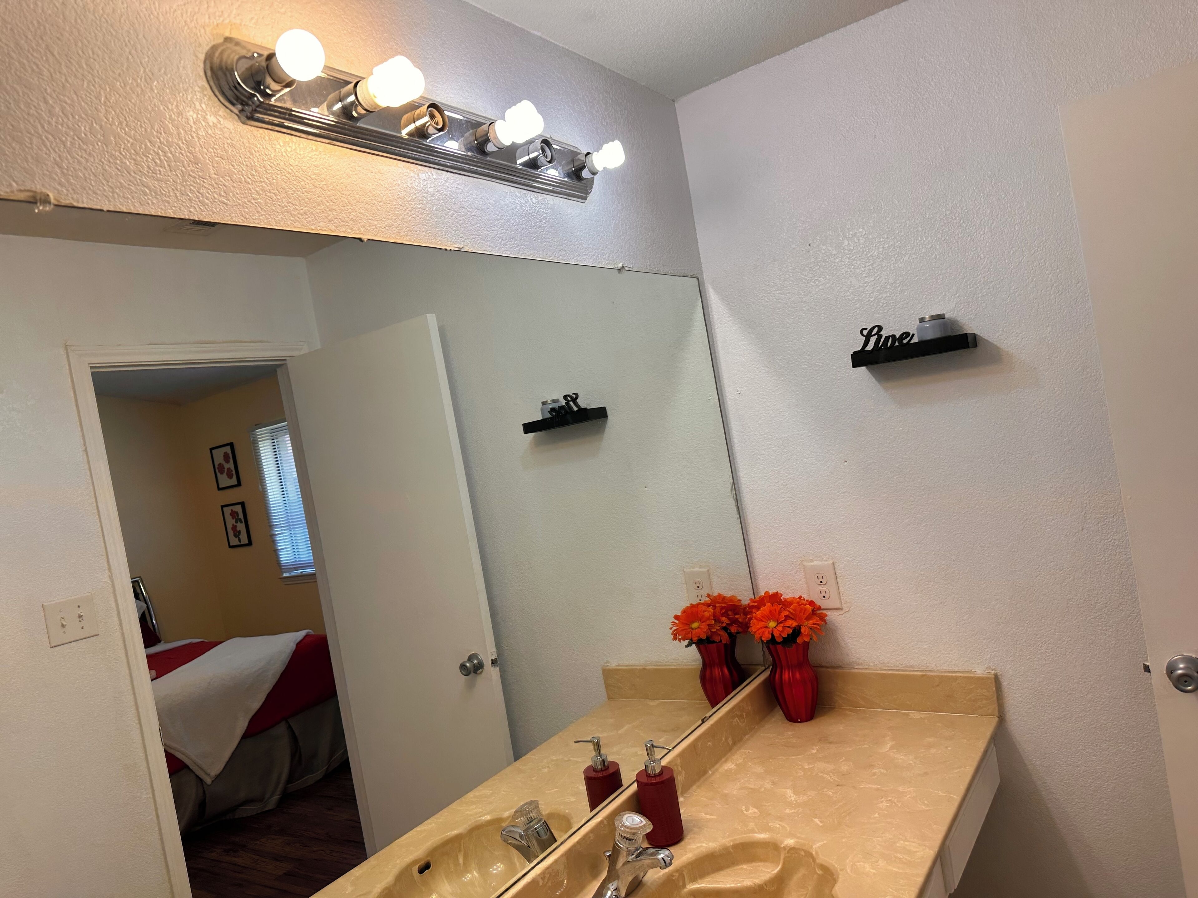 Un bagno con un lavandino color beige, uno specchio e un mobile contenitore con un vaso di fiori rossi. Ci sono delle luci sopra lo specchio e uno scaffale sopra il lavandino.