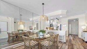 A modern kitchen with white cabinetry, a central island, and a dining area with wicker chairs and a glass table. - Seachase 303E: Gulf Views & Premium Amenities (Orange Beach)