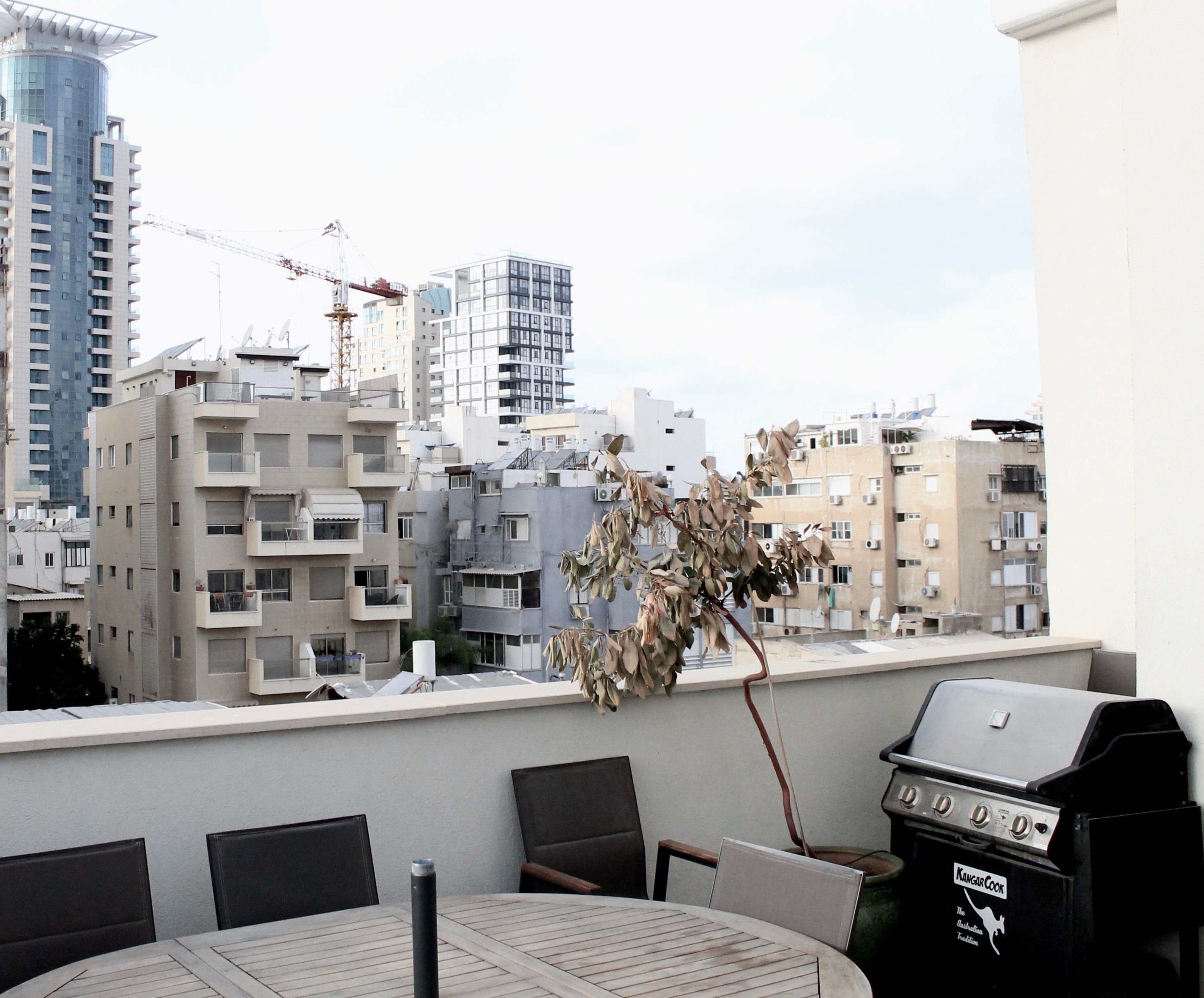 A balcony with a grill and chairs, overlooking a cityscape with multiple buildings and a construction crane.
