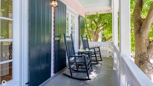 Une véranda avec deux fauteuils à bascule et une table, une balustrade blanche et un arbre en arrière-plan.