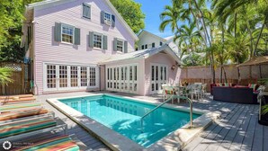 Una casa de dos pisos con piscina, mobiliario exterior y palmeras.