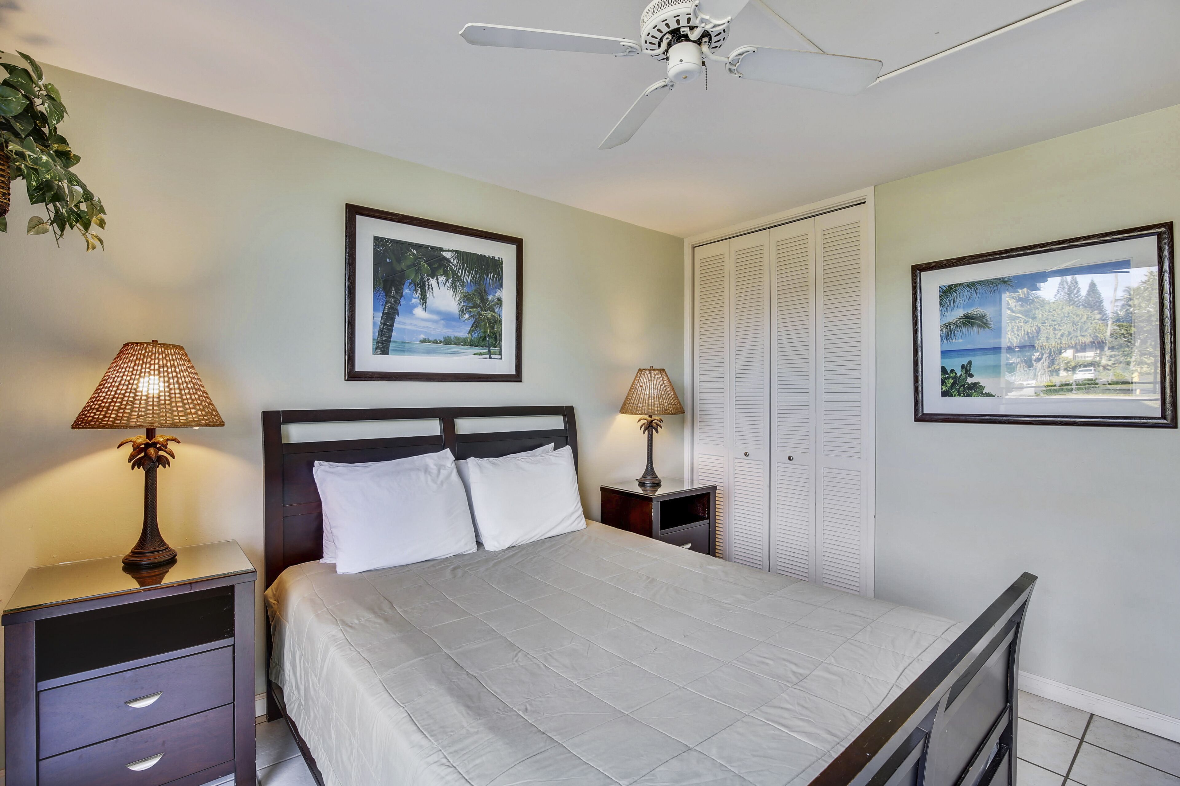 A bedroom with a bed, two bedside tables with lamps, a ceiling fan, and two framed pictures on the walls.