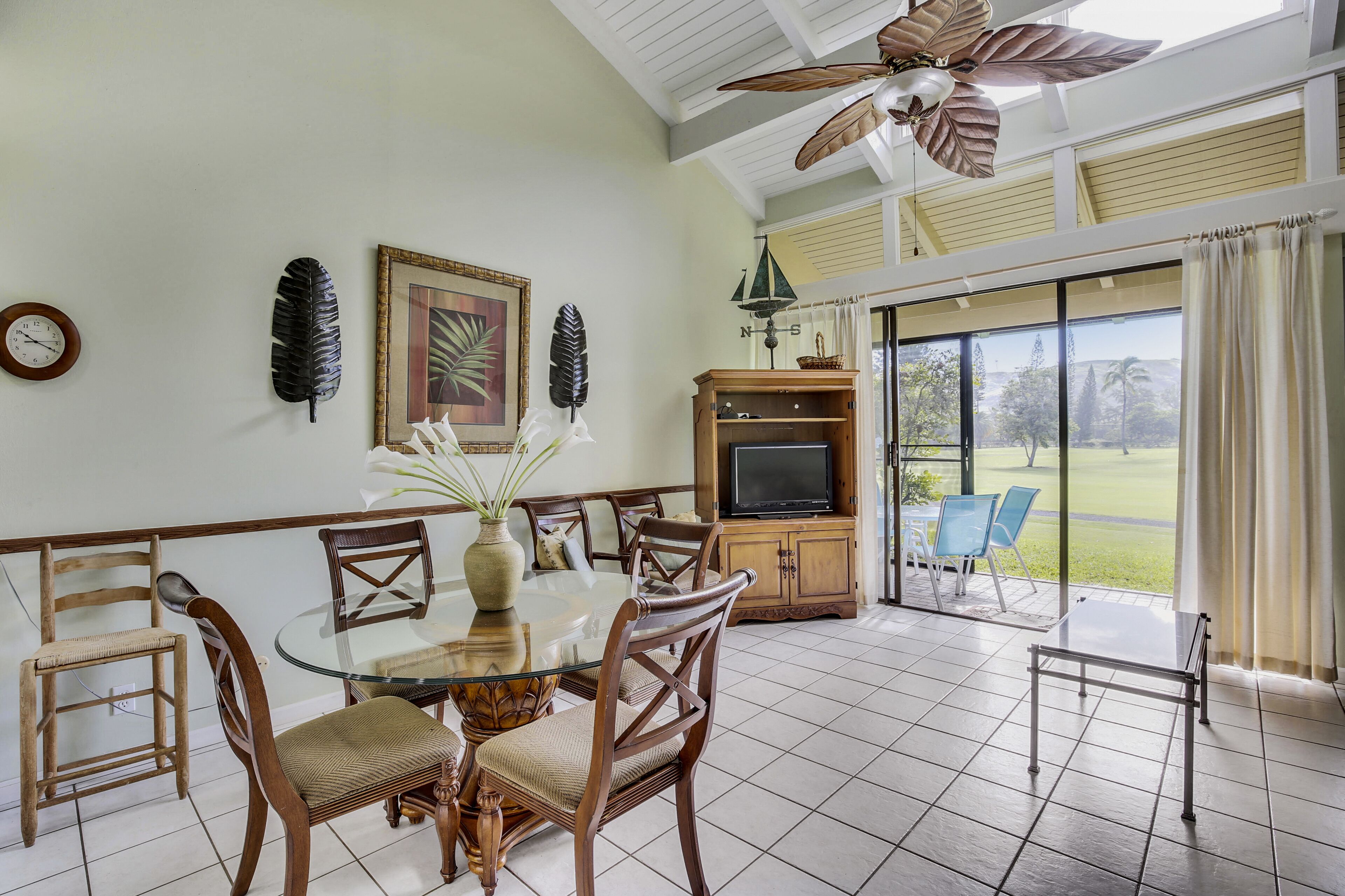 A dining area with a glass table, wooden chairs, a television, and a view of a golf course through large windows.