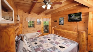 Une chambre dans un chalet en bois, avec un lit, un ventilateur de plafond, une télévision et une fenêtre avec des rideaux.