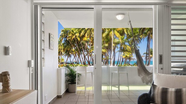 A modern living room with a sliding glass door leading to a balcony with a view of palm trees and the sea.