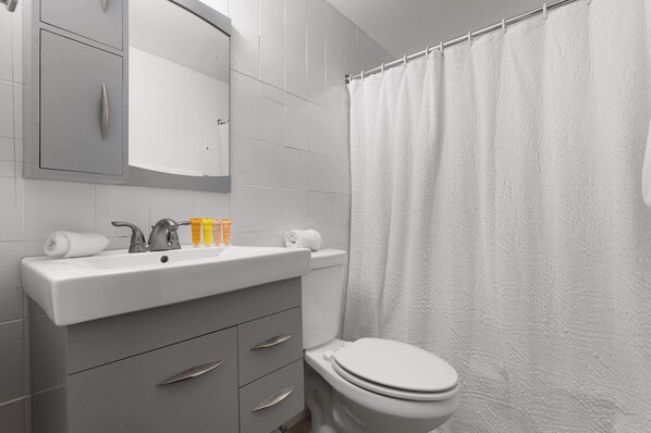 Un baño moderno con un lavamanos blanco, espejo y retrete. Hay artículos de aseo en el lavamanos y una cortina de ducha.