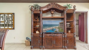A wooden entertainment center with a flat-screen TV displaying a resort view, flanked by decorative shelves and a mirror. - Sonoran Sun E-708 Sunset Serenity Relaxing Oceanfront Condo (Puerto Penasco)