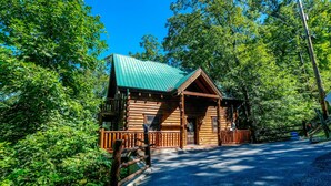 Une cabane en rondins avec un toit végétalisé, une terrasse en bois et une allée bordée d’une clôture.
