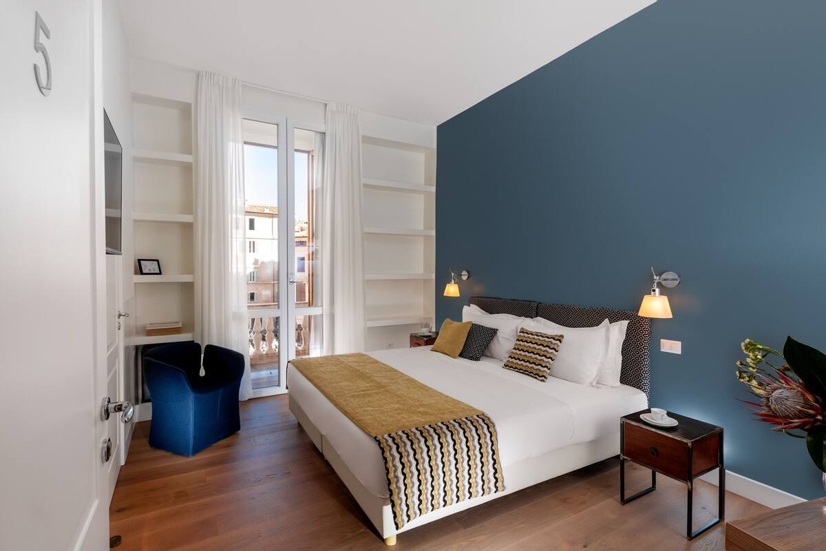 A bedroom with a large bed, a nightstand, a blue chair, and a view of the outside.