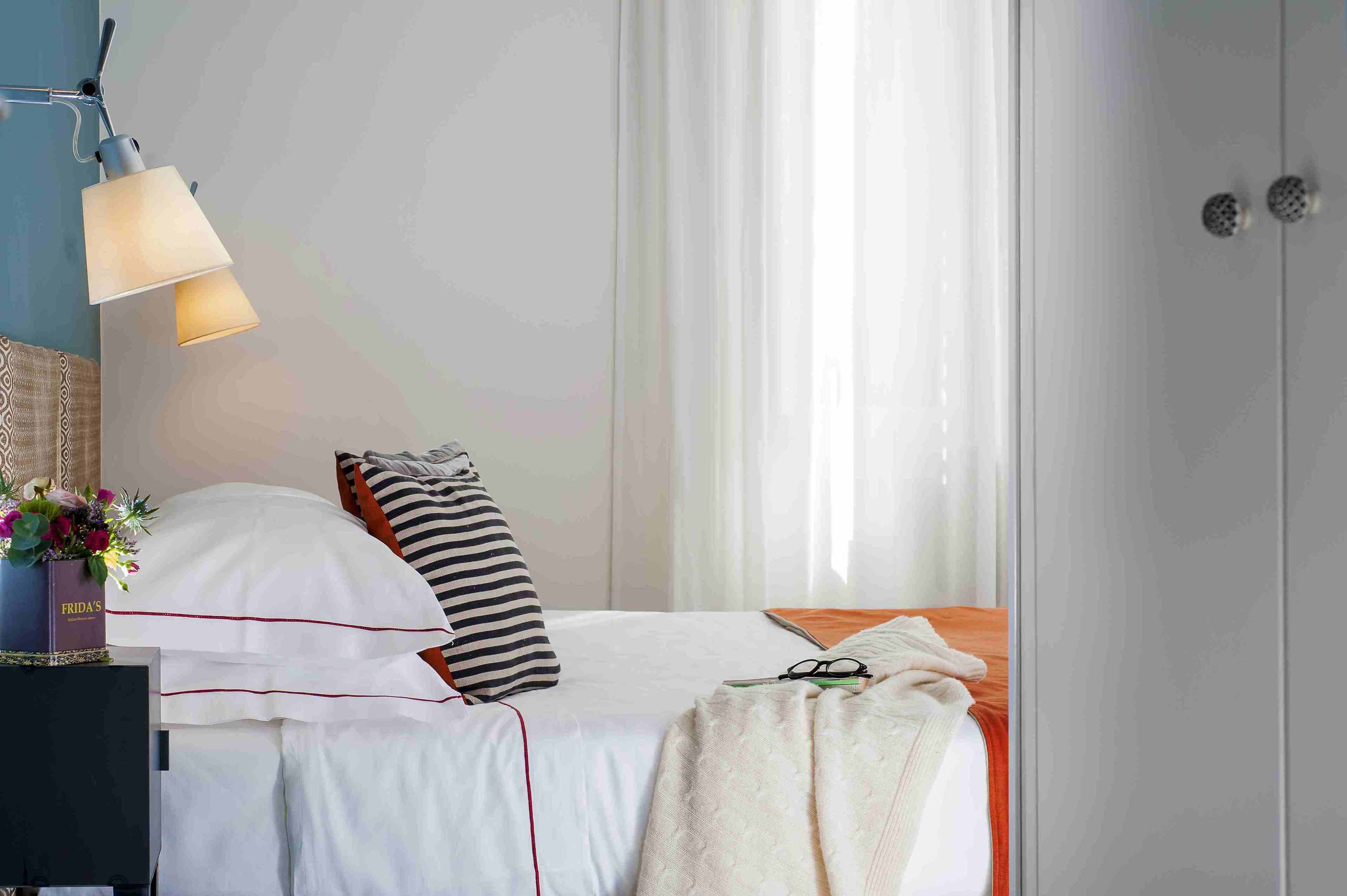 Ein ordentlich bezogenes Bett mit weißer Bettwäsche und gestreiften Kissen, ein Nachttisch mit Lampe und ein Fenster mit durchscheinenden Vorhängen.