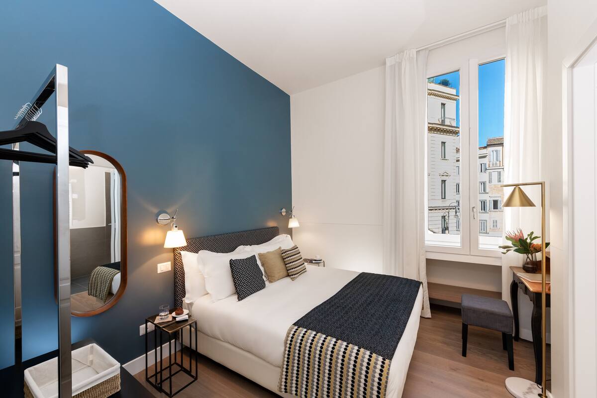 A modern hotel room with a large bed, a desk, a chair, a mirror, and a window with a city view.