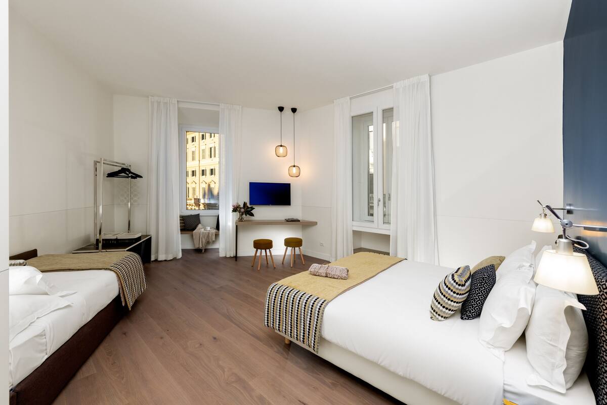 A hotel room with two beds, a desk, a TV, and a view of buildings.