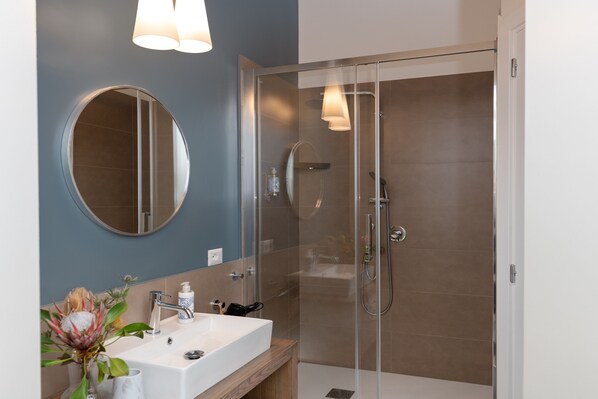 Une salle de bain moderne avec une douche à parois de verre, un miroir rond et un lavabo blanc avec un meuble-vasque en bois.