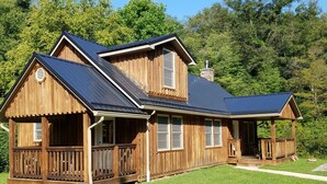 Eine Holzhütte mit Veranda und Schornstein, umgeben von einem grünen Rasen und Bäumen.