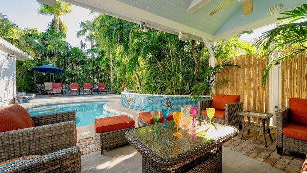 Zona junto a la piscina con mobiliario de mimbre, una piscina con azulejos y abundante vegetación.
