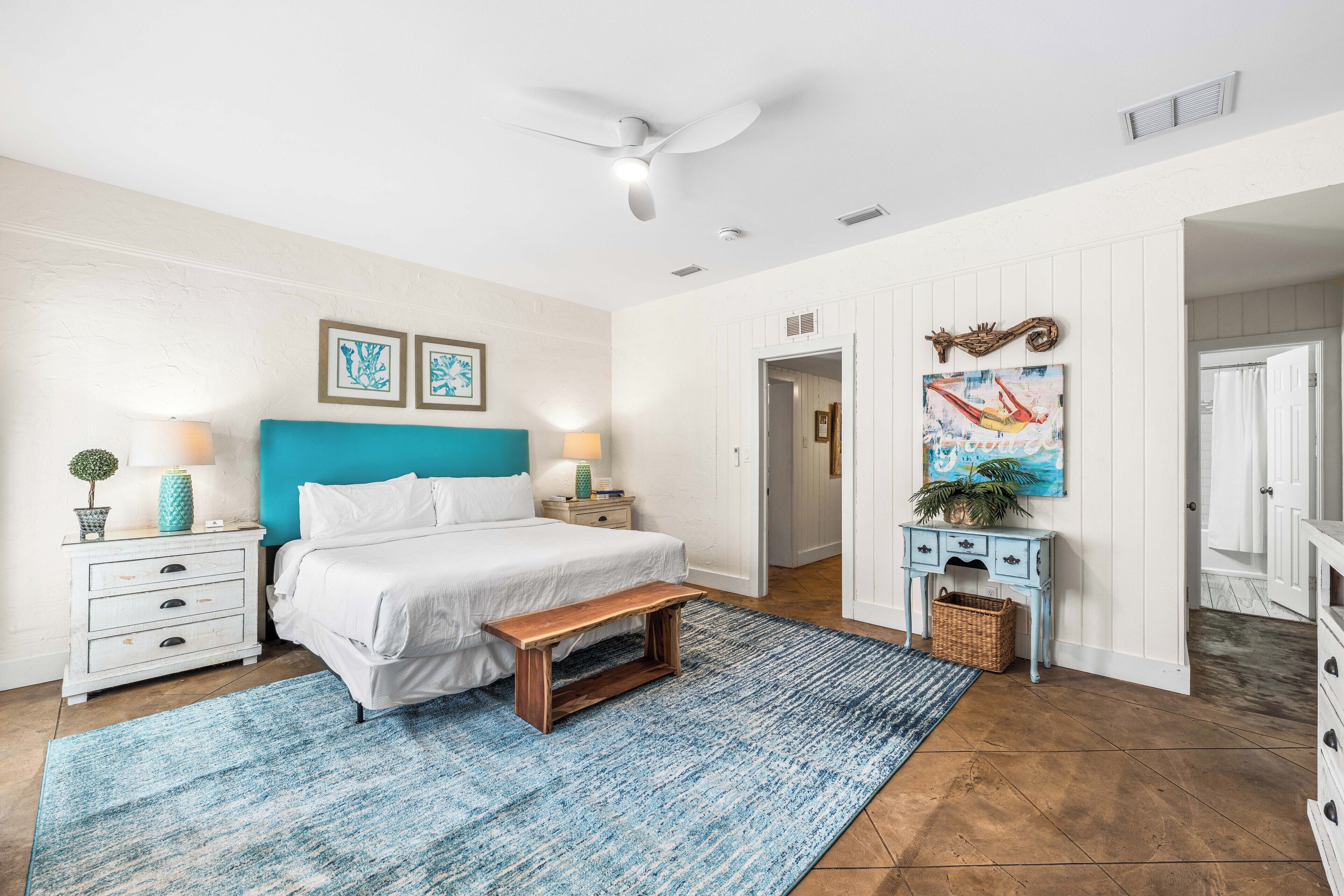 Un dormitorio con cama, banqueta, mesita de noche, lámpara, un cuadro en la pared y ventilador de techo.