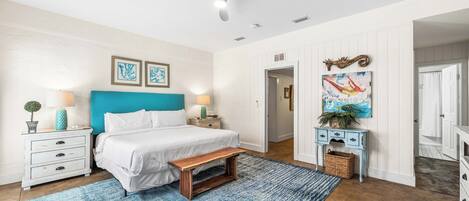 Un dormitorio con cama, banqueta, mesita de noche, lámpara, un cuadro en la pared y ventilador de techo.