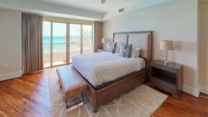 A spacious bedroom with a large bed, a bench, a nightstand, and a view of the ocean through large windows. - Astonishing Oceanfront Residence Located at The Ritz-Carlton, Grand Cayman (Seven Mile Beach)