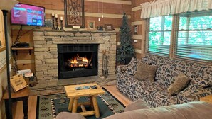 A cozy living room with a stone fireplace, a television, a patterned sofa, and a Christmas tree. - 1BR log cabin (sleeps 4) with screened-in hot tub and 1 free Dollywood ticket! (Sevierville)