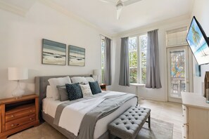 A bedroom with a bed, bedside table, lamp, artwork, window with curtains, and a television.