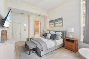 Quarto com cama, mesa de cabeceira, banqueta e TV fixada na parede.