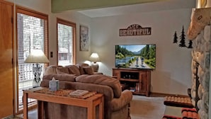 Ein Wohnzimmer mit einer Couch, einem Fernseher, einem Tisch mit einer Lampe und einer Wand mit dem Schriftzug 'Life is Beautiful'.