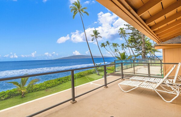 A balcony with a view of the ocean, palm trees, and a lounge chair. - Superior, remodeled, Oceanfront two bedroom 2.5 bath low rise, AC, pool. (Lahaina)