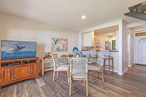 A dining area with a wooden table and wicker chairs, a television on a wooden cabinet, and a kitchen in the background. - Superior, remodeled, Oceanfront two bedroom 2.5 bath low rise, AC, pool. (Lahaina)