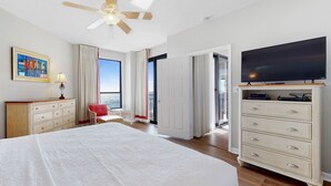 A hotel room with a large bed, a ceiling fan, a TV on a dresser, a desk with a chair, and a view of the sea through the window. - Leave your worries behind in this Relaxing Gulf Condo! - Recently Updated! (Orange Beach)