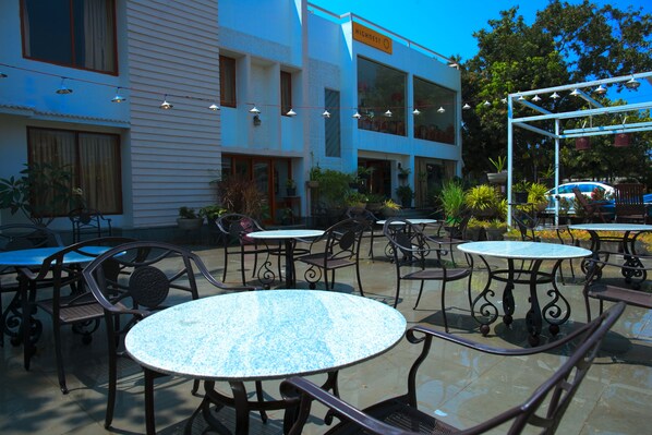 Un patio al aire libre con mesas y sillas redondas, rodeado de plantas en macetas y un edificio con un letrero que dice "HIGHEST 3".