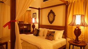 Quarto com cama de dossel, cabeceira de madeira e um relógio de parede grande. Há uma mesa de cabeceira com uma luminária e uma cadeira ao fundo.