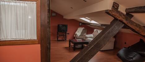 A loft-style living room with a sloped ceiling, wooden beams, and a large window with sheer curtains.