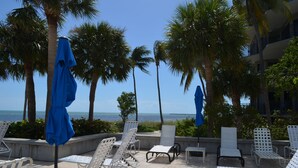 A poolside area with white metal chairs and blue umbrellas, palm trees, and a view of the ocean. - Botanical Island Delight  Key West fantastic discounts for Summer & Fall (Key West)