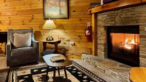 A cozy log cabin interior with a stone fireplace, a television mounted above it, a comfortable armchair, a small table with a lamp, and a framed picture on the wall.