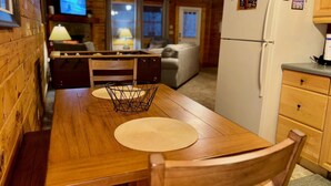 A wooden interior with a dining table, chairs, a kitchen area with a refrigerator, and a television in the background.