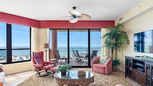 Ein geräumiges Wohnzimmer mit großem Fenster und Blick auf einen Strand, ausgestattet mit einer Couch, Sesseln, einem Couchtisch und einem Fernseher.