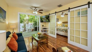 Ein gemütliches Wohnzimmer mit Sofa, Couchtisch und Bücherregal. Eine Glastür öffnet sich zu einem Balkon mit Blick auf Grünfläche.