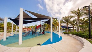 Un parque infantil con un tobogán y estructuras para escalar, rodeado de palmeras y cubierto por un toldo con sombra.
