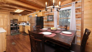 Interno di una baita in legno con un tavolo da pranzo imbandito per quattro persone, pavimento in legno, cucina con elettrodomestici moderni e un'ampia finestra con vista sugli alberi.