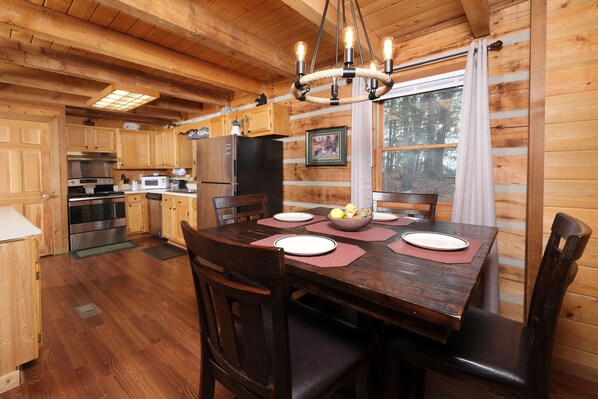Interior de una cabaña de madera con una mesa de comedor para cuatro, pisos de madera, una cocina con electrodomésticos modernos y un ventanal con vistas a árboles.