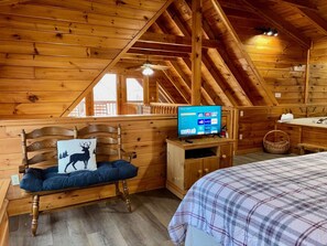 A wooden cabin interior with a bed, a bench with a deer pillow, a TV on a wooden stand, and a bathroom with a sink and a basket.