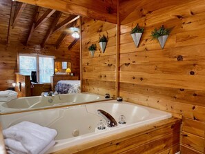 A wooden cabin interior with a large bathtub, wooden walls, and a mirror reflecting the room. - Pet friendly, hot tub, cozy log cabin sleeps 7! (Sevierville)